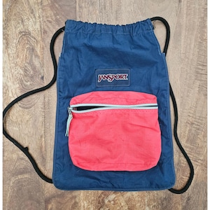 May include: A navy blue Jansport drawstring backpack with a red front pocket. The backpack has a black drawstring and the Jansport logo is visible on the front. The pocket has a zipper closure.
