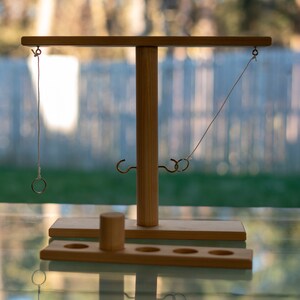 May include: A wooden ring toss game with a central vertical post and a horizontal bar. Two metal hooks and two rings are visible. The game includes a wooden base with holes and a small wooden cylinder. The background is blurred.
