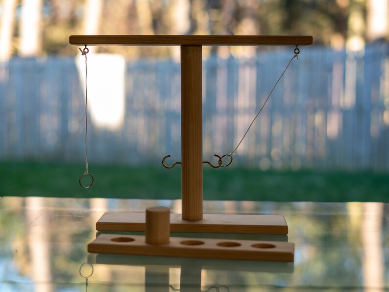 May include: A wooden ring toss game with a central vertical post and a horizontal bar. Two metal hooks and two rings are visible. The game includes a wooden base with holes and a small wooden cylinder. The background is blurred.