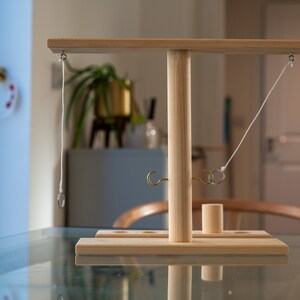 May include: A wooden ring toss game with a light-colored finish. The game features a T-shaped structure with a base, a central pole, and a horizontal bar. Rings are suspended from the bar, and hooks are attached to the base. A small wooden cylinder is also included.