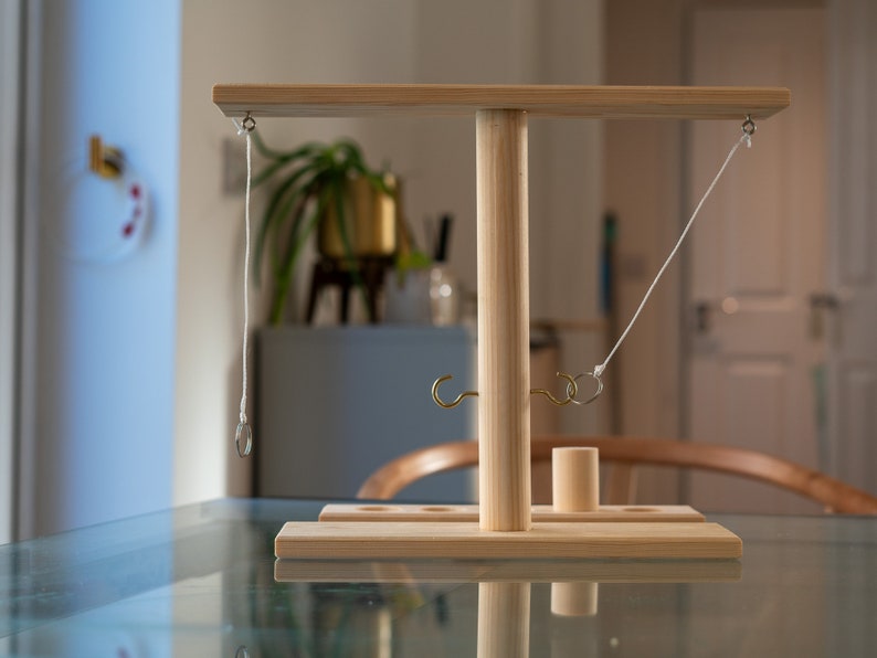 May include: A wooden ring toss game with a light-colored finish. The game features a T-shaped structure with a base, a central pole, and a horizontal bar. Rings are suspended from the bar, and hooks are attached to the base. A small wooden cylinder is also included.