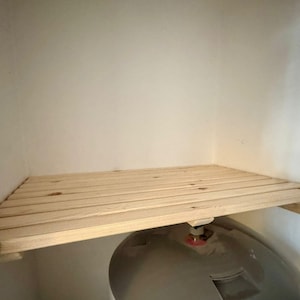 May include: A natural wood shelf, made from parallel wooden slats, is fixed to a wall. The shelf is supported by small wooden brackets. The background is a plain, off-white wall.