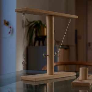 May include: A wooden ring toss game with a horizontal bar and a central vertical post. Two rings hang from strings attached to the bar. A small wooden base with a cylindrical peg sits nearby. The game is made of light-colored wood.