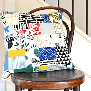 May include: A colourful patchwork cushion with geometric and floral patterns in shades of blue, red, yellow, black and white. The cushion is sitting on a wooden chair.
