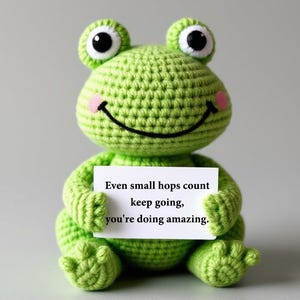 May include: A handmade green crochet frog with large black and white eyes and a smiling mouth. The frog holds a white sign that reads, "Even small hops count keep going, you're doing amazing."