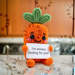 Pode incluir: Um brinquedo de cenoura de crochê com uma parte superior verde de folhas, segurando um cartão branco com o texto "Estou sempre torcendo por você!" A cenoura tem um rosto sorridente com olhos pretos e bochechas rosadas.