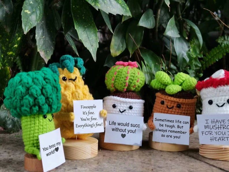 May include: Five crocheted plushies, each with a different design and a message. The plushies are a pineapple, a broccoli, a cactus, a succulent, and a mushroom. The messages are: "Fineapple It's fine. You're fine. Everything's fine!" "Life would succ without you!" "Sometimes life can be tough. But always remember so are you!" "I HAVE S MUSHRO FOR YOU HEART"