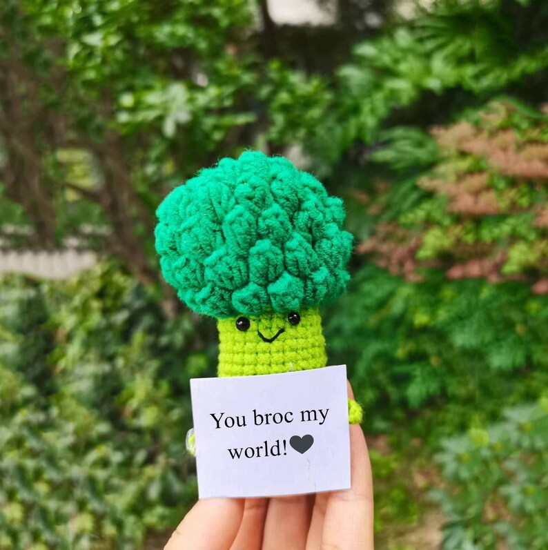 May include: A crocheted broccoli plush toy holding a card that says "You broc my world!" with a heart. The toy is green and has a happy face.