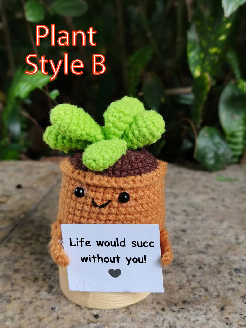 May include: A crocheted plant pot with a green plant and a sign that reads "Life would succ without you!" The pot has a smiling face and black eyes.