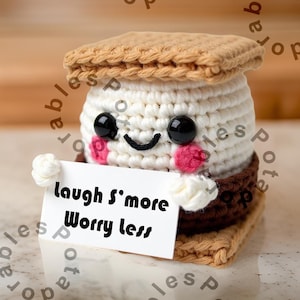 Puede incluir: Un peluche de s'more tejido a ganchillo con una cara sonriente, ojos negros y mejillas rosas. El s'more sostiene un letrero que dice "Laugh S'more Worry less". El peluche está hecho de hilo blanco, marrón y beige.
