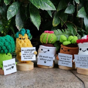 May include: Five crocheted plant characters holding signs with motivational messages. The characters are a pineapple, a cactus, a succulent, a broccoli, and a mushroom. The signs read: "be a pineapple", "Life would suck without you!", "Sometimes life can be tough. But always remember so are you!", "You broc my world!", and "I have something for you in my heart".