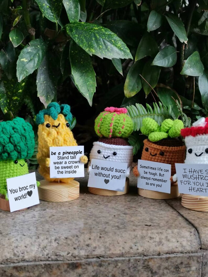 May include: Five crocheted plant characters holding signs with motivational messages. The characters are a pineapple, a cactus, a succulent, a broccoli, and a mushroom. The signs read: "be a pineapple", "Life would suck without you!", "Sometimes life can be tough. But always remember so are you!", "You broc my world!", and "I have something for you in my heart".