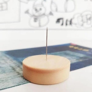 May include: A light brown, circular needle holder with a silver needle inserted in the center. The holder is resting on a blue and green patterned surface. The background features a white surface with a faint drawing.
