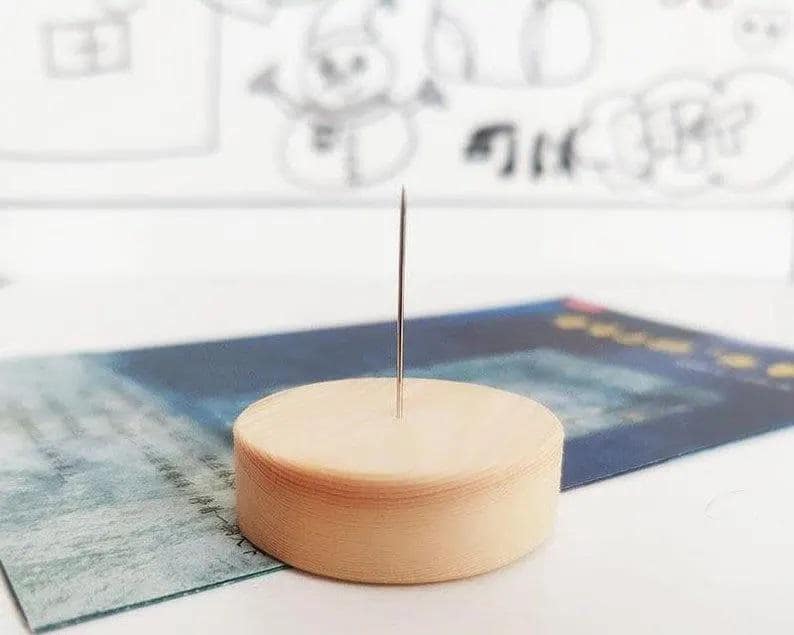 May include: A light brown, circular needle holder with a silver needle inserted in the center. The holder is resting on a blue and green patterned surface. The background features a white surface with a faint drawing.