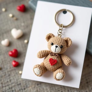May include: A brown crocheted teddy bear keychain with a red heart on its chest. The bear is wearing a brown bow tie and is attached to a gold key ring.