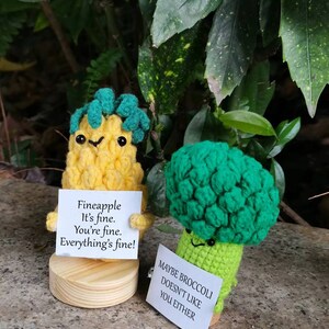 May include: Two crocheted plushies, one shaped like a pineapple and the other like a broccoli, holding signs with humorous messages. The pineapple plush has a yellow body, green leaves, and a sign that reads "Fineapple It's fine. You're fine. Everything's fine!" The broccoli plush has a green body and a sign that reads "Maybe broccoli doesn't like you either."