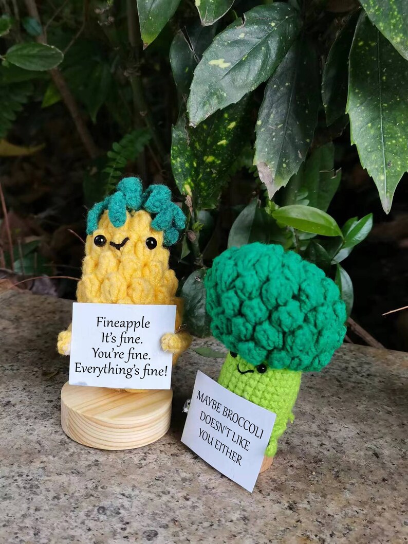 May include: Two crocheted plushies, one shaped like a pineapple and the other like a broccoli, holding signs with humorous messages. The pineapple plush has a yellow body, green leaves, and a sign that reads "Fineapple It's fine. You're fine. Everything's fine!" The broccoli plush has a green body and a sign that reads "Maybe broccoli doesn't like you either."