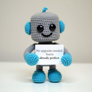 May include: A gray and blue crocheted robot toy with large black eyes and a small antenna on its head. The robot holds a white rectangular sign that features the black printed text: No upgrades needed. You're already perfect.