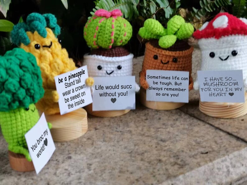 May include: Five crocheted amigurumi figures with different messages on signs. A green broccoli, a yellow pineapple, a green cactus, a brown succulent, and a white mushroom.