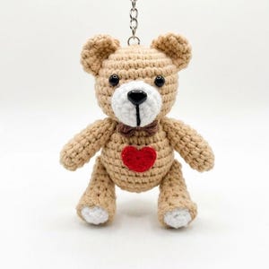Könnte beinhalten: Ein beigefarbener Häkel-Teddybär-Schlüsselanhänger mit weißer Schnauze, schwarzer Kunststoffnase, schwarzen Perlenaugen und einer kleinen braunen Fliege. Ein leuchtend rotes, gehäkeltes Herz ist auf die Brust genäht und die Füße haben weiße Akzente.