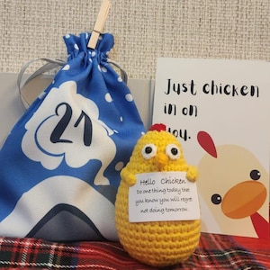 Op de afbeelding: Een geel gehaakt kippen speelgoed dat een wit kaartje vasthoudt met de tekst "Hello Chicken Do one thing today that you know you will regret not doing tomorrow." De kip zit op een rood-zwart geruit oppervlak naast een blauwe trekkoordtas met het nummer 21 erop en een wit kaartje met de tekst "Just chicken in on you."