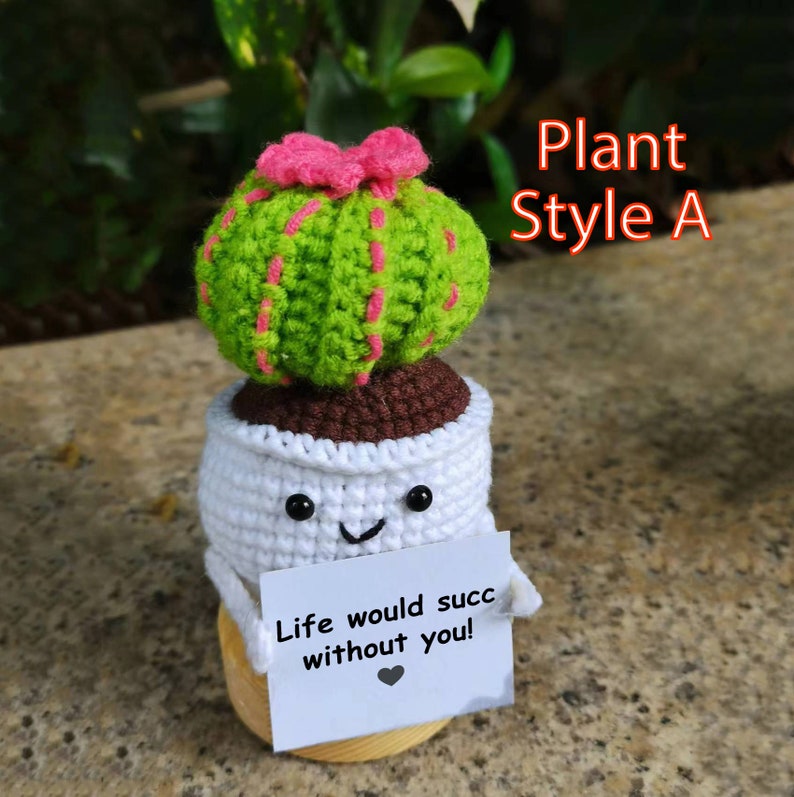 May include: A crocheted cactus plant in a white pot with a smiling face. The pot is sitting on a wooden base and has a card that says "Life would succ without you!"  Plant Style A.
