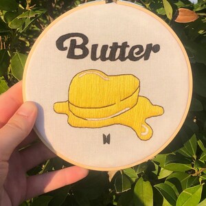Può includere: Un cerchio da ricamo bianco con la parola "Butter" ricamata in lettere corsive nere. Sotto la parola, è ricamato un disegno di burro giallo fuso. La lettera "V" è ricamata in nero sotto il disegno del burro.
