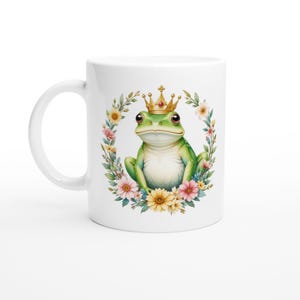 May include: White ceramic mug featuring a green frog wearing a gold crown and sitting in a wreath of colourful flowers.