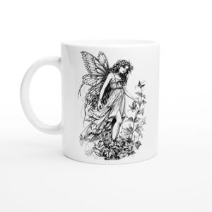 May include: White ceramic mug with a black and white illustration of a fairy with wings, a flower crown, and a flowing dress. The fairy is standing in a field of flowers.