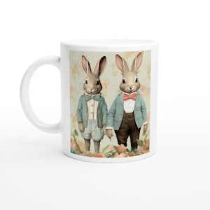 May include: White ceramic mug with a watercolour illustration of two rabbit figures in suits and bow ties. The rabbits are set against a floral backdrop. The mug has a white handle.