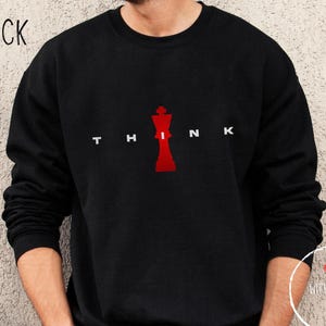 Può includere: Felpa nera a maniche lunghe con una grafica di re degli scacchi rossa e la parola "THINK" in lettere bianche.