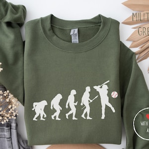 Op de afbeelding: Een groen sweatshirt met een witte afbeelding van de evolutie van honkbalspelers. De afbeelding bevat een honkbal en de tekst "With Love, Are".