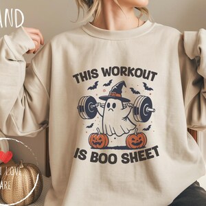 Puede incluir: Sudadera color arena con un gráfico de Halloween. El diseño presenta un fantasma con un sombrero de bruja levantando pesas, con calabazas y murciélagos. El texto dice "THIS WORKOUT IS BOO SHEET."