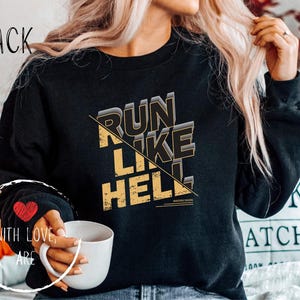 Könnte beinhalten: Schwarzes Sweatshirt mit dem Text "RUN LIKE HELL" in goldener und schwarzer Schrift. Das Wort "BLACK" ist oben links in Schreibschrift geschrieben. Ein rotes Herz ist auf dem linken Ärmel gestickt. Eine Person hält eine weiße Tasse.