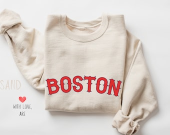 May include: A beige sweatshirt with the word "BOSTON" printed in red letters with a blue outline. The sweatshirt is folded in half and the text is centered on the front of the sweatshirt.