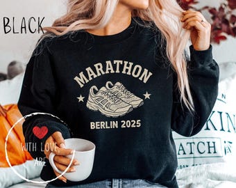 Könnte beinhalten: Schwarzes Sweatshirt mit dem Wort "MARATHON" über einer Grafik von Laufschuhen und dem Text "BERLIN 2025". Der Ärmel hat ein rotes Herz mit den Worten "WITH LOVE, ARE".