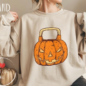 May include: Sand-colored sweatshirt featuring a Halloween-themed design. The graphic is an orange pumpkin with a handle, resembling a kettlebell. The pumpkin has carved-out eyes and a mouth. The word "SAND" is printed at the top left.