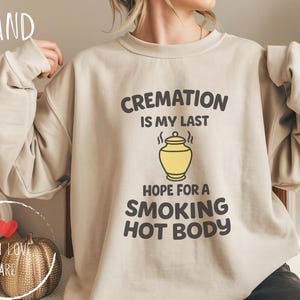 Puede incluir: Sudadera color arena con el texto negro "CREMATION IS MY LAST HOPE FOR A SMOKING HOT BODY". El diseño incluye un gráfico de urna amarilla. La palabra "SAND" está impresa en la esquina superior izquierda.