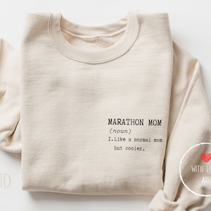 Puede incluir: Sudadera color arena con el texto negro "MARATHON MOM (sustantivo) I. Like a normal mom but cooler.". La sudadera tiene cuello redondo y mangas largas. Se ve un pequeño gráfico de corazón.