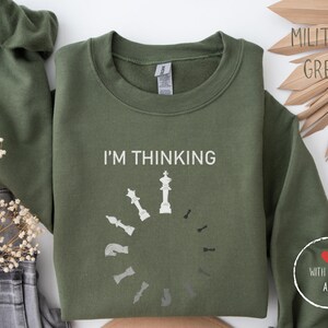 Può includere: Una felpa verde con la scritta "I'm thinking" e una grafica di una scacchiera con un re al centro. La grafica è un simbolo di caricamento con pezzi degli scacchi intorno al re. La felpa ha un cuore rosso con la scritta "With love, ARE" sulla manica.