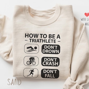 Puede incluir: Sudadera color arena con texto y gráficos en negro. El texto dice "HOW TO BE A - TRIATHLETE - DON'T DROWN, DON'T CRASH, DON'T FALL." También se ve un pequeño corazón rojo y las palabras "WITH LOVE, ARE".