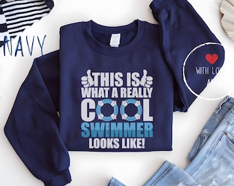 Op de afbeelding: Marineblauwe sweatshirt met de tekst "THIS IS WHAT A REALLY COOL SWIMMER LOOKS LIKE!" in wit en blauw. Het ontwerp bevat een duim omhoog en een reddingsboei-afbeelding. Een klein rood hart staat op de mouw.