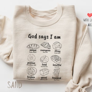 Może przedstawiać: Beżowa bluza z napisem "God says I am" i różnymi pozytywnymi afirmacjami, każda połączona z ilustracją o tematyce pieczywa. Bluza ma również nadrukowane słowa "SAND" i "WITH LOVE, ARE".