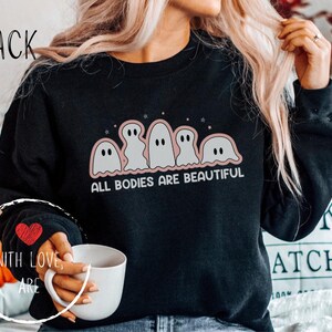 May include: Black sweatshirt with a graphic of five cartoon ghosts in a row, outlined in pink, with the text "ALL BODIES ARE BEAUTIFUL". The sleeve has a red heart. The word "BLACK" is at the top.