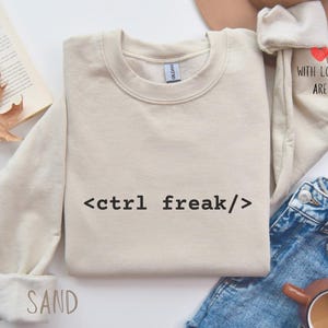 Può includere: Felpa color sabbia con la scritta "<ctrl freak/>" in nero. La felpa è abbinata a jeans blu e una tazza di caffè. La manica ha la scritta "WITH LOVE, ARE" e un cuore.