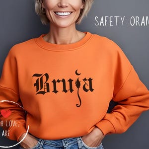 Puede incluir: Una sudadera naranja con la palabra "Bruja" en fuente gótica. La sudadera tiene cuello redondo y mangas largas. El texto "Safety Orange" está escrito encima de la sudadera. Un gráfico circular con las palabras "With Love, Are" está en la parte inferior izquierda.