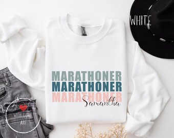 Könnte beinhalten: Weißer Pullover mit dem dreimal wiederholten Text "MARATHONER" in einem Farbverlauf von Blau, Grün und Rosa. Der Name "Samantha" ist in Schwarz unter dem Text geschrieben.
