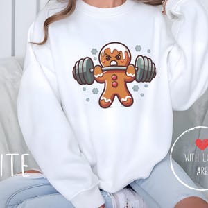 Könnte beinhalten: Weißes Sweatshirt mit einem Cartoon-Lebkuchenmann, der Gewichte hebt. Der Lebkuchenmann hat einen wütenden Gesichtsausdruck und ist mit weißem Zuckerguss verziert. Auf dem Sweatshirt steht das Wort "WHITE".