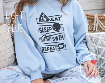 Op de afbeelding: Lichtblauw sweatshirt met de zwarte woorden "EAT SLEEP SWIM REPEAT". Het woord "EAT" staat boven een pizzapunt, "SLEEP" boven een slapend gezicht, "SWIM" boven een zwemmer en "REPEAT" heeft een cirkelpijl.