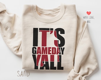 May include: A cream-colored sweatshirt with the text "It's Gameday Y'all" in black and red lettering. The red lettering is shaped like the state of Alabama.